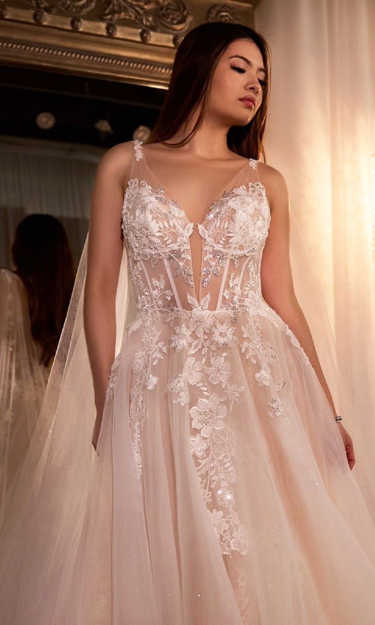 A woman wearing the Ladivine CDS476W Long Wedding Dress, a sleeveless sheer lace gown with floral appliqués, stands indoors in front of a mirror.